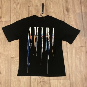 Svart AMIRI t-shirt med färgstänk - Svart t-shirt från AMIRI med stort tryck på ryggen och mindre tryck på bröstet. Trycket består av AMIRI-loggan med färgstänk i blått, vitt och orange som rinner ner. Klassisk passform och rund hals. Perfekt för dig som gillar streetwear med edge.