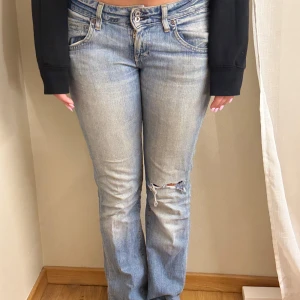 Ljusblå bootcut jeans med slitning - Säljer ett par ljusblå jeans med bootcut passform och slitning på ena knät. Jeansen har klassisk femficksdesign, låg midja och är tillverkade i denim. Perfekta för en avslappnad och trendig look. Lite slitna ner till
