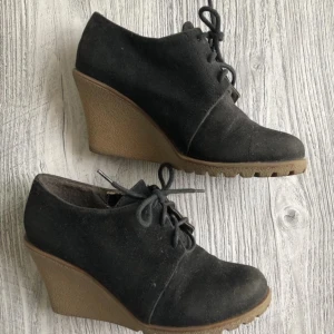 Svarta kilklackar från Rebel 36 - Snygga svarta kilklackar från Rebel, storlek 36 i mocka med snörning framtill. Skorna har en stabil brun kilklack och grov sula för extra grepp. Perfekta för dig som vill ha både höjd och komfort i vardagen.