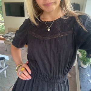 Svart kort klänning med spetsdetaljer - Supersöt svart kort klänning med resår i midjan och volangkant nertill. Klänningen har korta ärmar, spetsdetaljer och diskret struktur i tyget. Perfekt för dig som gillar en feminin och chill stil.