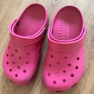 Rosa Crocs sandaler - Säljer ett par klassiska Crocs i en stark rosa färg. Skorna har ventilerande hål, justerbar hälrem och är tillverkade i lätt syntetmaterial. Perfekta för sommardagar eller när du vill ha något bekvämt och färgglatt på fötterna.