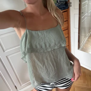 Ljusgrön linnetopp med volang - Supersöt ljusgrön linnetopp med tunna axelband och volangdetalj framtill. Toppen har en luftig och flowy passform, samt öppen rygg med djup urringning. Perfekt för varma dagar och ger en chill vibe till din outfit.