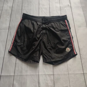 Svarta Moncler badshorts med ränder Boxer Mare beach shorts - You can buy this only on Vinted profile: brysu1 Svarta badshorts från Moncler med röda och vita ränder längs sidorna och klassisk Moncler-logga på vänster ben. Shortsen har elastisk midja och är tillverkade i lätt polyester, perfekta för sommaren och strandhäng.