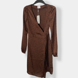 Brun omlottklänning med leopardmönster - Snygg brun omlottklänning från Vila med svart leopardmönster. Klänningen har långa ärmar, v-ringning och knyt i midjan för en smickrande passform. Perfekt för dig som vill sticka ut med ett trendigt djurmönster.