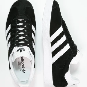 Adidas Gazelle svart/vit mockasneaker - Klassiska Adidas Gazelle sneakers i svart mocka med vita detaljer och de ikoniska tre ränderna på sidorna. Skon har snörning, vit platt sula och guldtexten 'GAZELLE' på sidan. Perfekt för dig som gillar retrostil och streetwear. Dom e använda men om man tvättar dom sk dom se ut som nya dom har Inge defekter förutom smutsiga.