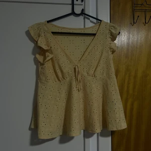En Gul blus med volang och broderi - Supersöt gul blus med broderade hålmönster och volangdetaljer på axlarna. Djup v-ringning med knyt framtill ger en romantisk vibe. Perfekt för varma dagar och passar till både jeans och kjol. 💛