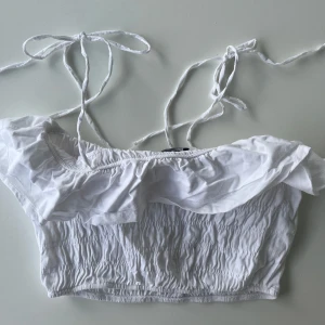Vit volang topp från Zara - Supersöt vit croppad topp från Zara med volangdetalj upptill och smockad midja. Axelbanden knyts på axlarna. Storlek M men passar även S.