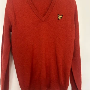 Roströd v-ringad tröja från Lyle & Scott - Säljer en roströd v-ringad tröja från Lyle & Scott med deras klassiska gula fågel-logga på bröstet. Tröjan är stickad och har långa ärmar samt ribbade muddar. Perfekt för dig som gillar stilren och tidlös design. Stl M