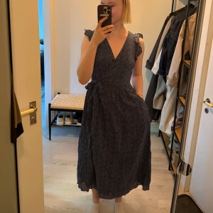 Blommig omlottklänning & Other Stories - Lång, marinblå omlottklänning från & Other Stories Paris Atelier med små vita blommor, volangdetaljer vid axlarna och knytband i midjan. Perfekt för dig som gillar flowiga och feminina siluetter.