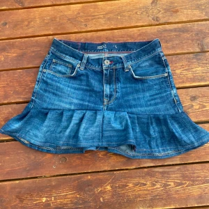 Jeanskjol med volang från Tommy Hilfiger - Snygg blå jeanskjol från Tommy Hilfiger med volang nertill och klassiska fickor fram och bak. Kjolen har bälteshällor och knappstängning framtill. Perfekt för dig som vill ha en cool och avslappnad stil. Jag har sytt den själv, men allt är noggrant gjort💕 den har en slitning mellan bakfickorna vilket syns på bild, men skulle inte säga att det stör💗Vet inte exakt storlek, men kan skicka mått vid intresse💗. Jag har flera liknade kjolar till salu!🫶🏻