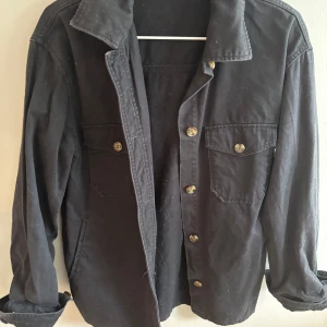 Svart overshirt från H&M i bomull - Svart overshirt från H&M i bomull med klassisk krage, knäppning framtill och två bröstfickor med knappar. Perfekt lager-på-lager-plagg med avslappnad passform och långa ärmar. Enkel och stilren design som funkar till det mesta.
