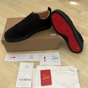 Christian Louboutin svarta sneakers med nitar - Exklusiva svarta sneakers från Christian Louboutin med ikonisk röd sula och svarta nitar på tån. Skorna har snörning, rund tå och är tillverkade i skinn och mocka. Perfekt för dig som vill sticka ut med en lyxig och edgy stil.