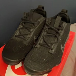 Nike Vapormax svarta sneakers - Svarta Nike Vapormax sneakers med stickad ovandel . Skorna har snörning och den ikoniska genomskinliga bubbliga sulan. Perfekta för dig som gillar streetstyle och vill ha något bekvämt och snyggt.