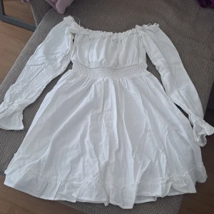 Vit offshoulder kortklänning med volang - Superfin vit kortklänning med offshoulder och långa ärmar. Klänningen har smock i midjan för en snygg passform och volangdetaljer längs nederkanten. Perfekt för sommarens alla tillfällen och ger en romantisk vibe.