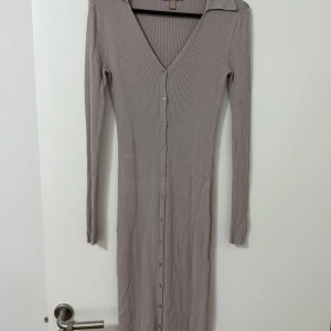 Lång ribbad  klänning med knappar - Snygg långklänning i ribbad beige tyg med v-ringning och långa ärmar. Klänningen har knappar hela vägen framtill och en klassisk krage. Den är figurnära och passar perfekt för dig som gillar en stilren look. I gott skick från H&M