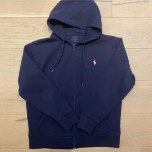 Marinblå hoodie från Polo Ralph Lauren - Säljer en marinblå hoodie från Polo Ralph Lauren med dragkedja och huva. Tröjan har snörning vid huvan, två fickor framtill och den klassiska vita loggan broderad på bröstet. Perfekt för chill dagar och enkel att matcha med jeans eller joggers.