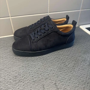  Christian Louboutin - Snygga svarta sneakers från Christian Louboutin i mocka med klassisk röd sula och svarta snören. Skorna har rund tå och platt sula, samt diskreta detaljer som ger en clean och lyxig vibe. Perfekta för dig som vill sticka ut med exklusiva skor.