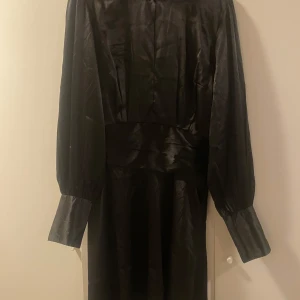 Svart klänning från Bubbleroom - Elegant svart klänning från Bubbleroom i glansigt material. Klänningen har långa ärmar med breda manschetter och markerad midja för en snygg siluett. Perfekt för dig som vill sticka ut med en stilren look.