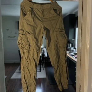 Beiga cargopants från H&M Divided - Säljer ett par beiga cargopants från H&M Divided med elastisk midja och flera stora fickor på sidorna. Byxorna är i bomull och har en avslappnad passform, perfekta för en chill och streetig stil.