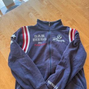 Blå zip-jacka från North Sails (retro) - Mörkblå zip-tröja från North Sails med broderad text 'San Diego' och 'Racer' framtill. Röda och vita ränder på axlarna, Storlek L men passar S-L enligt mig. Köpt för länge sedan och är en retro tröja tror den kosta runt nutidens 1800kr.  Sparsamt använd. Tveka inte att höra av er vid frågor och intresse!