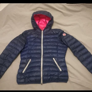 Blå dunjacka med rosa foder från Colmar - Snygg blå pufferjacka från Colmar med rosa foder och huva. Jackan har dragkedja framtill och två snedställda fickor med dragkedja. Perfekt för kyliga dagar och ger en sportig look. Jackan är använd ett fåtal gånger.  Ny priset är 3500:-
