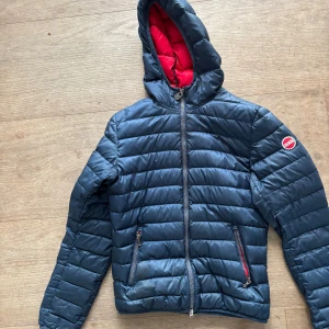 Mörkblå pufferjacka med röd huva - Säljer en mörkblå pufferjacka med röd insida på huvan. Jackan har dragkedja framtill och två fickor med röda dragkedjor. Perfekt för kyligare dagar och har en sportig look. För barn strl 14år/166cm. Använd men bra skick