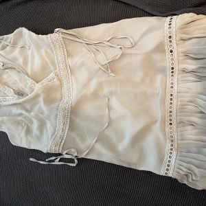 Ljusgrå omlottklänning från AllSaints - Supersöt ljusgrå omlottklänning från AllSaints med v-ringning och fina spetsdetaljer. Klänningen har knytband i midjan och volang nertill, samt dekorativa broderade kanter. Perfekt för sommarens alla tillfällen!