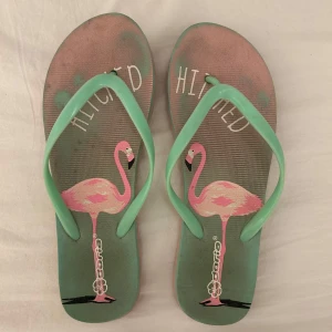 Gröna flip-flops med flamingo från Doria - Säljer ett par gröna flip-flops med mintgröna remmar och ett tryck av rosa flamingos på sulan. Perfekta för sommaren och strandhäng. Från Märket Doria i strl 38