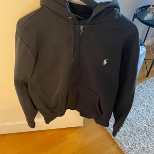 Svart hoodie med dragkedja från Polo Ralph Lauren - Klassisk svart hoodie från Polo Ralph Lauren med dragkedja och huva. Tröjan har två fickor framtill och den ikoniska broderade loggan i vitt på bröstet. Perfekt för en avslappnad stil. Bra skick på tröjan 8/10