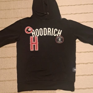 Svart hoodie från Hoodrich med broderade detaljer - Svart hoodie från Hoodrich med broderade märken och text på bröstet. Tröjan har huva med dragsko och en stor ficka framtill. Snygga röda och vita detaljer som ger en sportig look.
