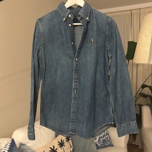 Jeansskjorta från Ralph Lauren - Säljer en klassisk blå jeansskjorta från Ralph Lauren med button-down krage och broderad logga på bröstet. Skjortan har långa ärmar och knäppning framtill. Perfekt för en avslappnad men stilren look.