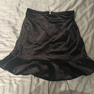 Svart volangkjol från H&M Divided - Säljer en svart kjol från H&M Divided i storlek 38. Kjolen har en snygg volang nertill och dragkedja bak. Materialet är glansigt och känns mjukt mot huden. Perfekt för dig som vill ha en stilren men ändå trendig look.