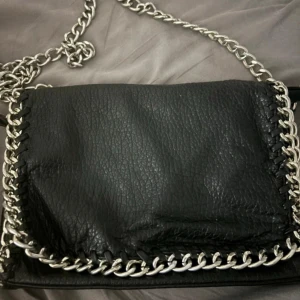 Svart axelväska med kedjedetaljer från Tiamo - Säljer en svart axelväska från Tiamo i skinnimitation med grova silverfärgade kedjor längs kanterna. Pris kan sänkas vid snabb affär