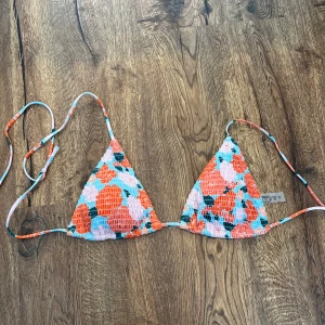 Blommig trekantsbikini med knyt - Säljer en färgglad trekantsbikini med blommigt mönster i orange, rött, blått och vitt. Bikinin har tunna knytband i sidorna och runt nacken, perfekt för sommardagar på stranden.
