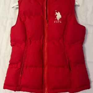 Röd dunväst från U.S. Polo Assn. - Röd dunväst från U.S. Polo Assn. med broderad logga på bröstet och dragkedja framtill. Västen är vadderad och har en sportig look, perfekt för lager-på-lager-stil. Tillverkad i polyester.