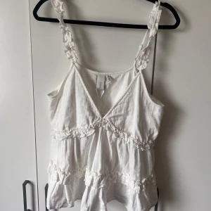 Vit volangprydd linnetopp från YAS - Supersöt vit linnetopp från YAS med volangdetaljer och smala axelband. Toppen har v-ringning och dekorativas rynkade band längs både fram- och baksida. 