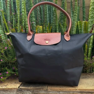 Svart handväska från Longchamp - Säljer en klassisk svart handväska från Longchamp med bruna handtag och lock i skinn. Väskan har guldknapp och är rymlig med enkel design, perfekt för dig som gillar stilrena accessoarer.