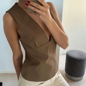Brun ärmlös topp från Mango - Säljer en snygg brun ärmlös topp från Mango med djup v-ringning och figurnära passform. Toppen har stilren design och är tillverkad i ett slätt material som ger en elegant look. Perfekt att matcha med jeans eller kjol.