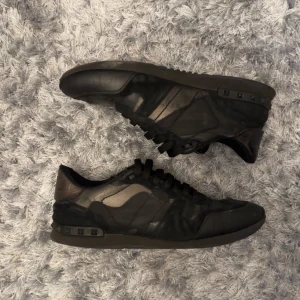 Valentino Rockrunner sneakers i svart och grått - Säljer ett par Valentino Rockrunner sneakers i svart och grått med diskret camouflage-mönster. Skorna har snörning, platt sula och klassiska nitar på hälen. Perfekta för dig som gillar exklusiva och stilrena sneakers.