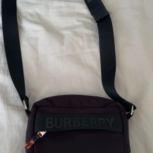Svart axelväska från Burberry - Säljer en svart axelväska från Burberry med justerbart axelband och tydlig logga framtill. Väskan har en dragkedja upptill och en stilren design som passar till många tillfällen.möts i Norrköping 