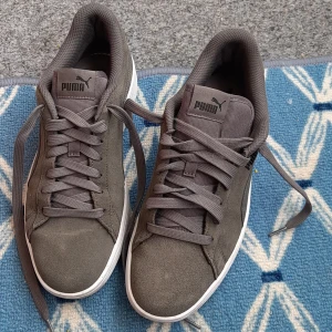 Grå sneakers från Puma - Säljer ett par stilrena grå sneakers från Puma med vit sula. Skorna har klassisk snörning och är tillverkade i mocka med diskret Puma-logga på sidan och plös. Perfekta för dig som gillar en enkel och sportig look. Skor är i mycket bra skick. 
