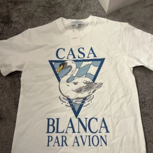Vit t-shirt med svan-tryck från Casablanca - Vit t-shirt från Casablanca med stort blått tryck framtill av en svan och texten 'Casa Blanca Par Avion'. Klassisk passform och rund halsringning. Perfekt för dig som gillar unika prints och stilrena plagg.