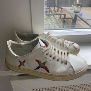 Vita sneakers från Axel Arigato med broderade fåglar - Säljer ett par vita sneakers från Axel Arigato med broderade fåglar i rött, blått, grönt och beige på sidorna och tån. Skorna har snörning och platt sula. Perfekta för dig som gillar stilrena men unika detaljer. Nypris 2750kr