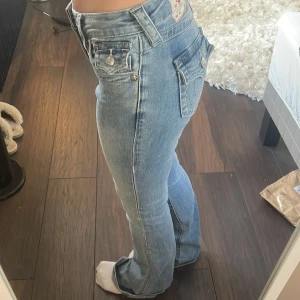 True Religion Becca Bootcut Jeans - Säljer ett par ljusblå jeans från True Religion, modell Becca Mid Rise Bootcut. Jeansen har klassiska fickor med lock och knappar, samt bootcut-ben. Snygga detaljer och ikonisk True Religion-lapp bak. 