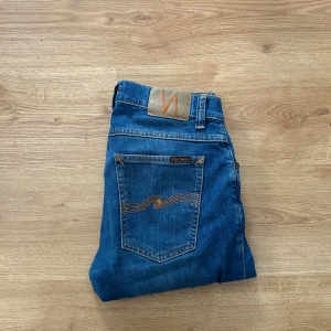 Blå jeans från Nudie Jeans - Säljer ett par klassiska blå jeans från Nudie Jeans med orangea sömmar och broderad bakficka. Modellen har normal passform och raka ben. Perfekta för dig som gillar stilrena och tidlösa jeans.