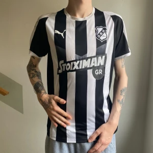 Fotbollströja Puma OFI Crete - En svartvit randig fotbollströja från Puma med OFI Crete FC-logga ⚽️ och Stoiximan sponsortryck på bröstet. Skön på sommaren. Perfekt om man ska vara målis eller domare om du är på det humöret.