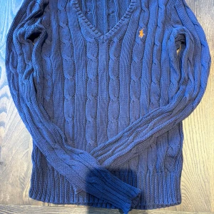 Blå stickad v-ringad tröja från Ralph Lauren - Mörkblå stickad tröja från Ralph Lauren med v-ringning och klassiskt kabelstickat mönster. Tröjan har långa ärmar och en broderad orange logga på bröstet. Nypris är 1800kr. Den är i väldigt fint skick och har bara använt några fåtal gånger. Skriv till mig om ni har några frågor!