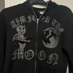 Svart hoodie Aim For The Moon med strassmotiv - Svart hoodie med dragkedja från Aim For The Moon. Framsidan har ett stort strassmotiv med en tecknad djävul och måne samt texten 'Aim For The Moon'. Huva och känguruficka framtill.