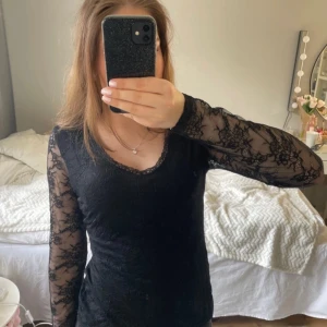 Svart spetsklänning med lång ärm - Säljer en svart klänning med långärmade spetsärmar och v-ringning. Klänningen har en tight passform och är dekorerad med blommigt spetsmönster över hela ärmarna och axelpartiet. Perfekt för dig som gillar en elegant och stilren look.