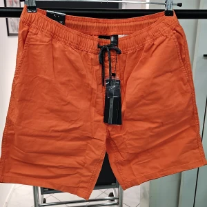 Orange shorts från O'Neill - Säljer ett par orange shorts från O'Neill med svart snörning i midjan och elastisk resår. Shortsen har sidofickor och är tillverkade i ett lätt syntetmaterial, perfekta för stranden eller poolen. Vintage effekt. Material/ Bomull.